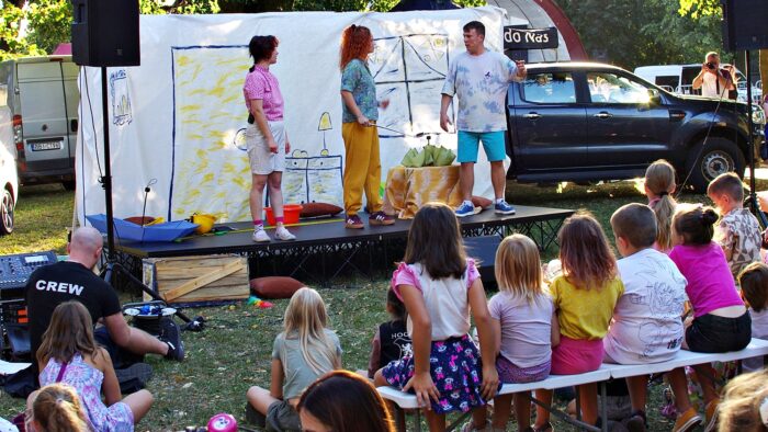 Plenerowy spektakl teatru. Małą scena w parku, przed nią ławki i widzowie.