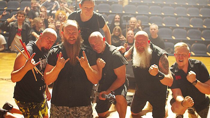 Zespół rockowy na scenie pozuje do fotografii. W tle - widownia pełna fanów.