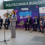 Kobieta przemawia do mikrofonu. Za nią siedzą pozostali uczestnicy konferencji.