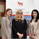 Trzy kobiety pozują do zdjęcia na tle roll-upu z herbem miasta.