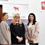 Trzy kobiety pozują do zdjęcia na tle roll-upu z herbem miasta.
