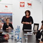 Losowanie. Dwie kobiety na szczycie długiego stołu. Jedna z nich mówi do osób siedzących po obu stronach stołu.