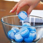 Losowanie. Biało-niebieskie kule w szklanym naczyniu. W kulach białe kartki. Nad naczyniem kobieca ręka z długimi paznokciami chwyta jedną kulę i wyjmuje ją z naczynia.