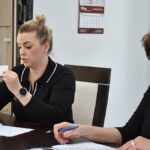 Dwie kobiety za stołem. Jedna czyta z kartki, druga pokazuje wylosowany numer, trzymając kartkę w dłoniach.