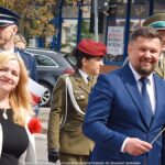 Uroczystości trzeciomajowe na terenie miasta. W przemarszu ulicami miasta idzie grupa osób. Na pierwszym planie uśmiechnięci: kobieta (z lewej - zastępca burmistrza) i mężczyzna (z prawej - burmistrz). W tle zabudowa miejska.