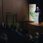 Mężczyzna stoi na scenie domu kultury, za mównicą i przemawia do zgromadzonych. Za nim widać fragment jasnego ekranu i roll-up. Przed nim ciemna część sali z widownią.