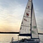 Jezioro i żaglówka z logotypami miasta i hufca na żaglu.