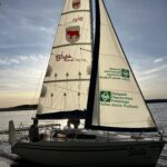 Jezioro i żaglówka z logotypami miasta i hufca na żaglu.