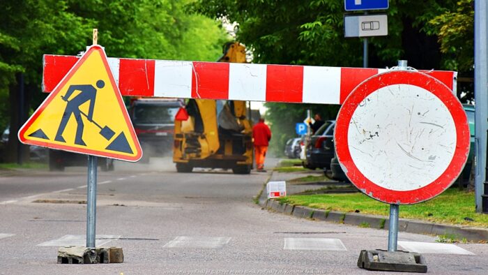 Droga zamknięta. Barierka w kolorach biało-czerwonych, na niej dwa znaki drogowe: roboty drogowe i zakaz ruchu, a w tle remontowana ulica i ekipa drogowców podczas pracy.