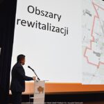 Widok ekranu na którym wyświetlona jest mapa i napis "Obszar rewitalizacji". Z lewej postać mężczyzny stojącego za mównicą i przemawiającego.