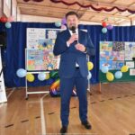 Burmistrz przemawiający do mikrofonu trzymanego w dłoni, podczas podsumowania konkursu. Wnętrze sali sportowej. Za burmistrzem prezentacja zwycięskich prac.