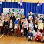 Grupowe zdjęcie dzieci - laureatów konkursu, ich opiekunów, organizatorów konkursu oraz gości rozdania nagród. W tle wystawa prac konkursowych oraz wnętrze sali sportowej.