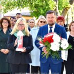 Grupowe zdjęcie burmistrzów i urzędników. W tle zieleń miejska. Na pierwszym planie mężczyzna trzyma bukiet biało-czerwonych kwiatów, a kobieta biało-czerwony znicz.