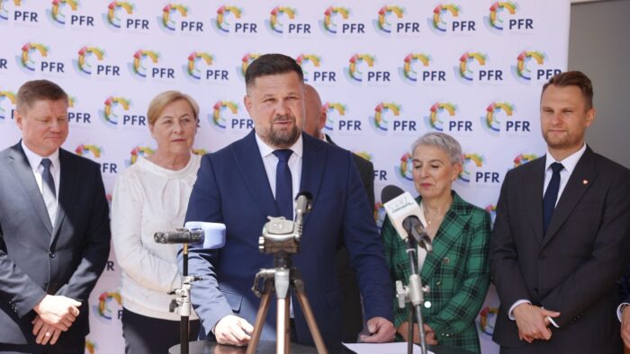 Konferencja prasowa w plenerze - na tle budynku biura. Do mikrofonu przemawia mężczyzna w garniturze - burmistrz. W tle pozostałe osoby uczestniczące w inauguracji biura.