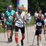 Zawody w nordic walkingu w lesie grabowieckim. Na pierwszym planie ścigają się trzej mężczyźni w równym wieku: od nastolatka po osobę starszą. W tle żwirowa droga, las, kibice i otoczenie wyścigu.