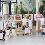 Wręczenie nagród w konkursie fotograficznym dla dzieci. Poziome ujęcie. Ustawione sztalugi z prezentowanymi pracami. Przy każdej z nich stoi młody autor fotografii. Po bokach stoją dwaj mężczyźni. Wszyscy pozują do wspólnego zdjęcia.