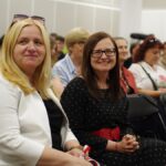 Dwie kobiety na widowni siedzą na krzesełkach i uśmiechają się, pozując do zdjęcia.