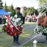 Mężczyzna - burmistrz niesie wieniec kwiatów, który ustawia przed mauzoleum na cmentarzu. Wokół otoczenie mauzoleum i trwające uroczystości.