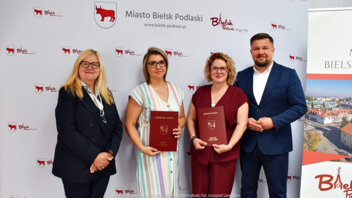 Na tle ścianki reklamowej miasta stoją: mężczyzna - burmistrz (z prawej) oraz trzy kobiety: zastępca burmistrza z lewej i dwie nowe dyrektorki. Wszyscy pozują do zdjęcia.