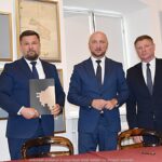 Trzej mężczyźni stoją w sali muzealnej i pozują do zdjęcia. Jeden z nich trzyma w rękach podpisaną umowę. W tle widać muzealną wystawę.