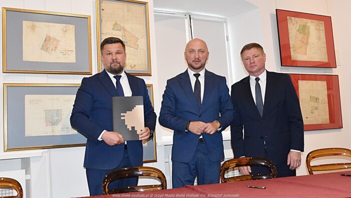 Trzej mężczyźni stoją w sali muzealnej i pozują do zdjęcia. Jeden z nich trzyma w rękach podpisaną umowę. W tle widać muzealną wystawę.