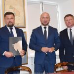 Trzej mężczyźni stoją w sali muzealnej i pozują do zdjęcia. Jeden z nich trzyma w rękach podpisaną umowę. W tle widać muzealną wystawę.