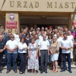 Zdjęcie grupowe kilkudziesięciu pracowników urzędu miasta przed budynkiem. Osoby stoją na schodach wejścia do urzędu i pozują do zdjęcia. Nad nimi złoty napis "urząd miasta".