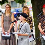 Uroczystość patriotyczna na placu przed ratuszem. Grupa młodych artystów, ustylizowana na powstańców, śpiewa stojąc pod drzewem. Z prawej - flaga Polski.