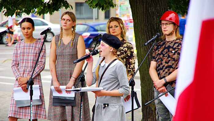 Uroczystość patriotyczna na placu przed ratuszem. Grupa młodych artystów, ustylizowana na powstańców, śpiewa stojąc pod drzewem. Z prawej - flaga Polski.