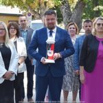 Grupa osób, pozuje do zdjęcia przed oddaniem hołdu przed pomnikiem. W środku mężczyzna trzyma znicz w barwach narodowych. W tle zieleń miejska.