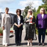 Sześć osób - mężczyzna i 5 kobiet - stoją w rzędzie na cmentarzu wojennym. Kobieta w środku kadru trzyma w rękach pionowy znicz z herbem miasta Bielsk Podlaski.