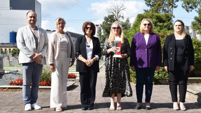 Sześć osób - mężczyzna i 5 kobiet - stoją w rzędzie na cmentarzu wojennym. Kobieta w środku kadru trzyma w rękach pionowy znicz z herbem miasta Bielsk Podlaski.