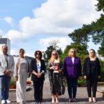 Sześć osób - mężczyzna i 5 kobiet - stoją w rzędzie na cmentarzu wojennym. Kobieta w środku kadru trzyma w rękach pionowy znicz z herbem miasta Bielsk Podlaski.