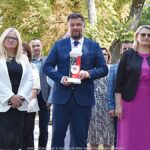 Grupa osób, pozuje do zdjęcia przed oddaniem hołdu przed pomnikiem. W środku mężczyzna trzyma znicz w barwach narodowych. W tle zieleń miejska.