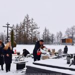 Członkowie delegacji miasta na zaśnieżonym cmentarzu, przed grobem, na którym składają wiązankę kwiatów i stawiają znicz.