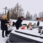 Członkowie delegacji miasta na zaśnieżonym cmentarzu, przed grobem, na którym składają wiązankę kwiatów i stawiają znicz.