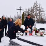 Członkowie delegacji miasta na zaśnieżonym cmentarzu, przed grobem, na którym składają wiązankę kwiatów i stawiają znicz.