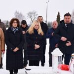 Członkowie delegacji miasta na zaśnieżonym cmentarzu, przed grobem, na którym składają wiązankę kwiatów i stawiają znicz.