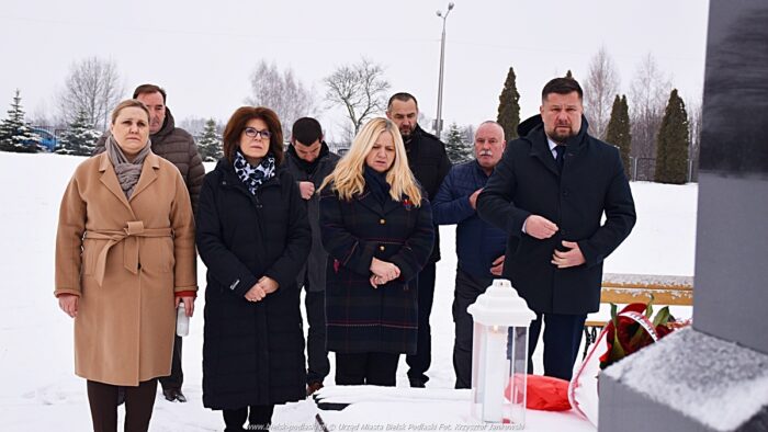 Członkowie delegacji miasta na zaśnieżonym cmentarzu, przed grobem, na którym składają wiązankę kwiatów i stawiają znicz.