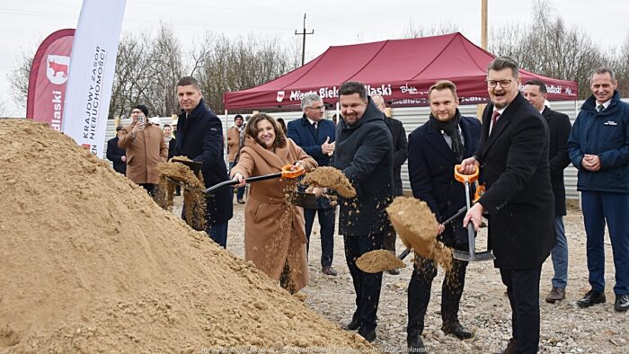Grupa oficjeli w uroczystych strojach z łopatami w dłoniach - przesypują piach na kupie.