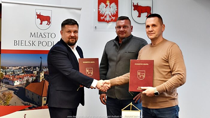 Trzech mężczyzn stoi i pozuje do zdjęcia. Dwaj mężczyźni po bokach prezentują dokumenty w okładkach miejskich i ściskają sobie dłonie. Przed nimi stoi stół, a na nich stojak z herbem miasta. W tle z lewej roll-up miejski, a powyżej na ścianie: godło Polski i herb miasta.