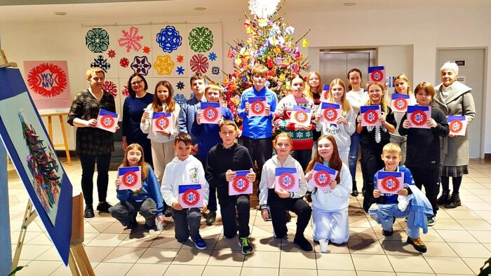 Grupowe zdjęcie uczestników zajęć z organizatorami. Dzieci trzymają w dłoniach grafiki. W tle kolorowa przystrojona choinka.