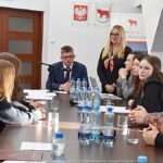 Obrady młodzieżowej rady miasta. Radni siedzą za stołem, z prawej stoi kobieta i do nich przemawia, z lewej operator z kamerą telewizyjną.