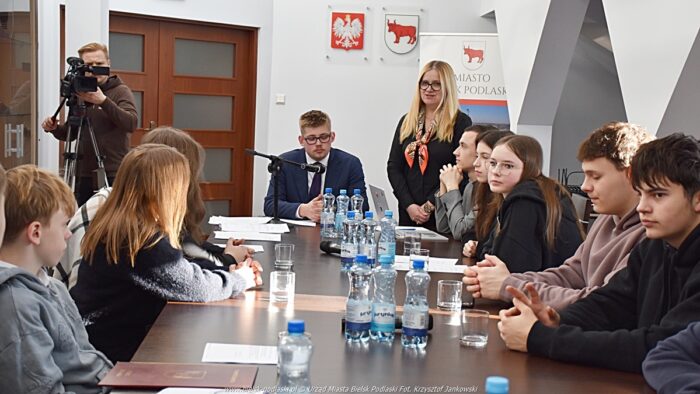 Obrady młodzieżowej rady miasta. Radni siedzą za stołem, z prawej stoi kobieta i do nich przemawia, z lewej operator z kamerą telewizyjną.