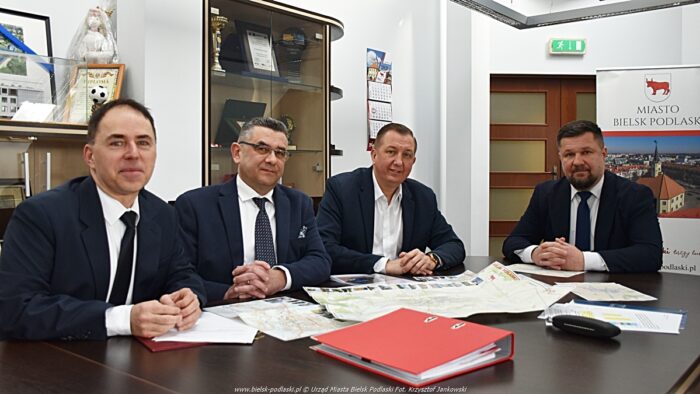 Czterej mężczyźni w garniturach siedzą za stołem w dużej sali konferencyjnej. Na stole rozłożone są dokumenty i mapy.