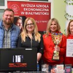 Wystawcy, młodzież i goście podczas prezentacji w liceum. Cztery osoby (trzy kobiety i mężczyzna) stoją na stoisku uczelni i pozują do zdjęcia z dyplomem i pucharem.