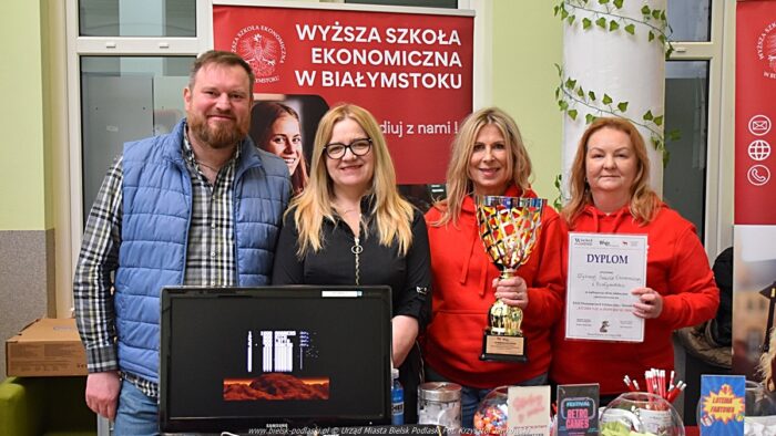 Wystawcy, młodzież i goście podczas prezentacji w liceum. Cztery osoby (trzy kobiety i mężczyzna) stoją na stoisku uczelni i pozują do zdjęcia z dyplomem i pucharem.