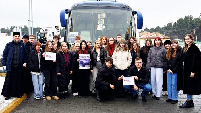 Zdjęcie kilkudziesięciu osób stojących na tle autokaru (w którym za szybą wisi proporczyk Bielskiego Klubu Sportowego Tur). Modzi ludzie trzymają w dłoniach plakat wyjazdu oraz logotypy "Bielsk Podlaski łączy ludzi".