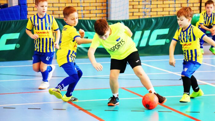Pięciu kilkulatków w strojach sportowych gra w piłkę na hali sportowej.