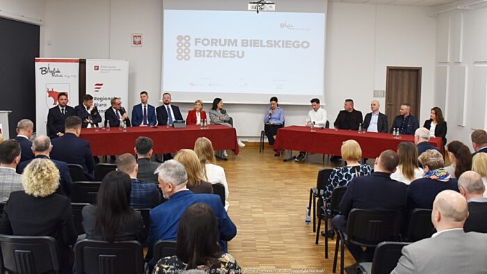 Szeroki plan na salę konferencyjną, wypełnioną gośćmi. Na środku kadru ekran z wyświetlonym napisem "Bielskie Forum Biznesu", pod nim dwa stoły po siedem osób, za którym siedzą dyskutujący prelegenci i dziennikarka prowadząca panel dyskusyjny, a przed nimi (tyłem) kilkudziesięciu przedsiębiorców siedzących na krzesłach i przysłuchujących się dyskusji.